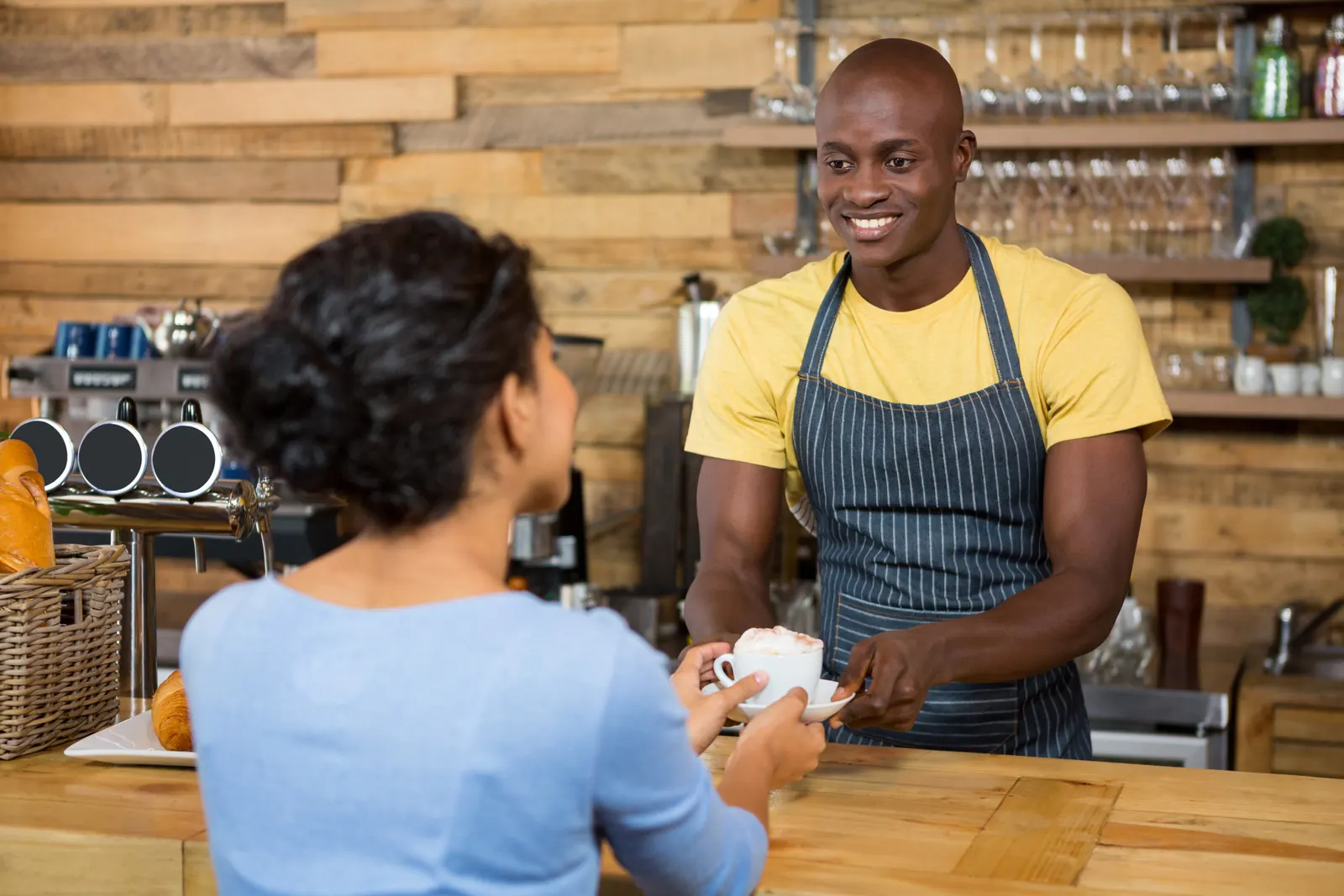 A smiling barista in a striped apron hands a cup of coffee across a wooden counter to a customer
