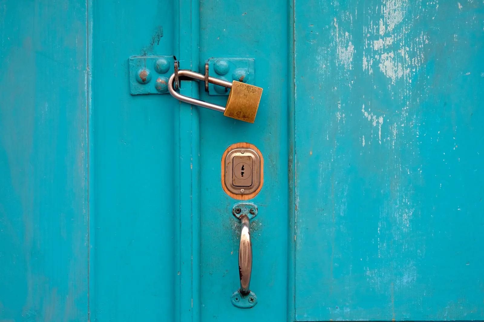 A brass padlock through the hasp of a weathered turquoise door, above a keyhole and a metal handle