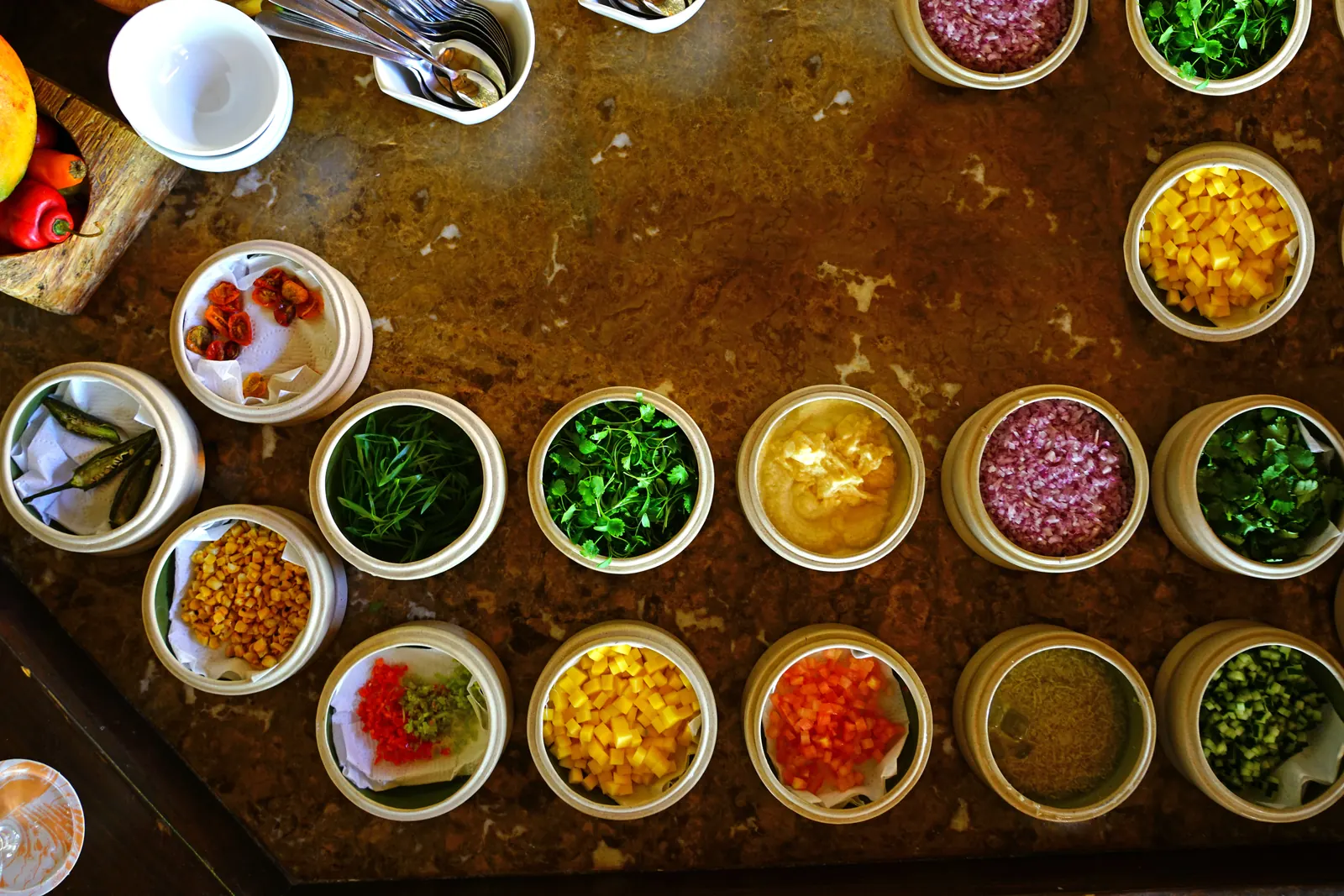 Rows of small white bowls of chopped herbs, vegetables and fruit laid out on a dark metal tray, seen from above