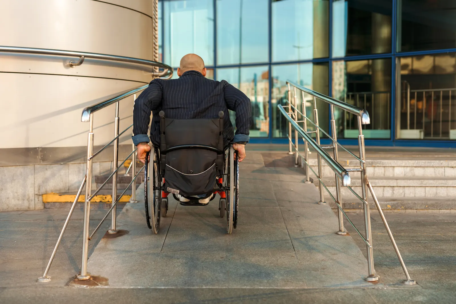 A man in a wheelchair, seen from behind, goes up a ramp with steel handrails toward a glass-fronted building